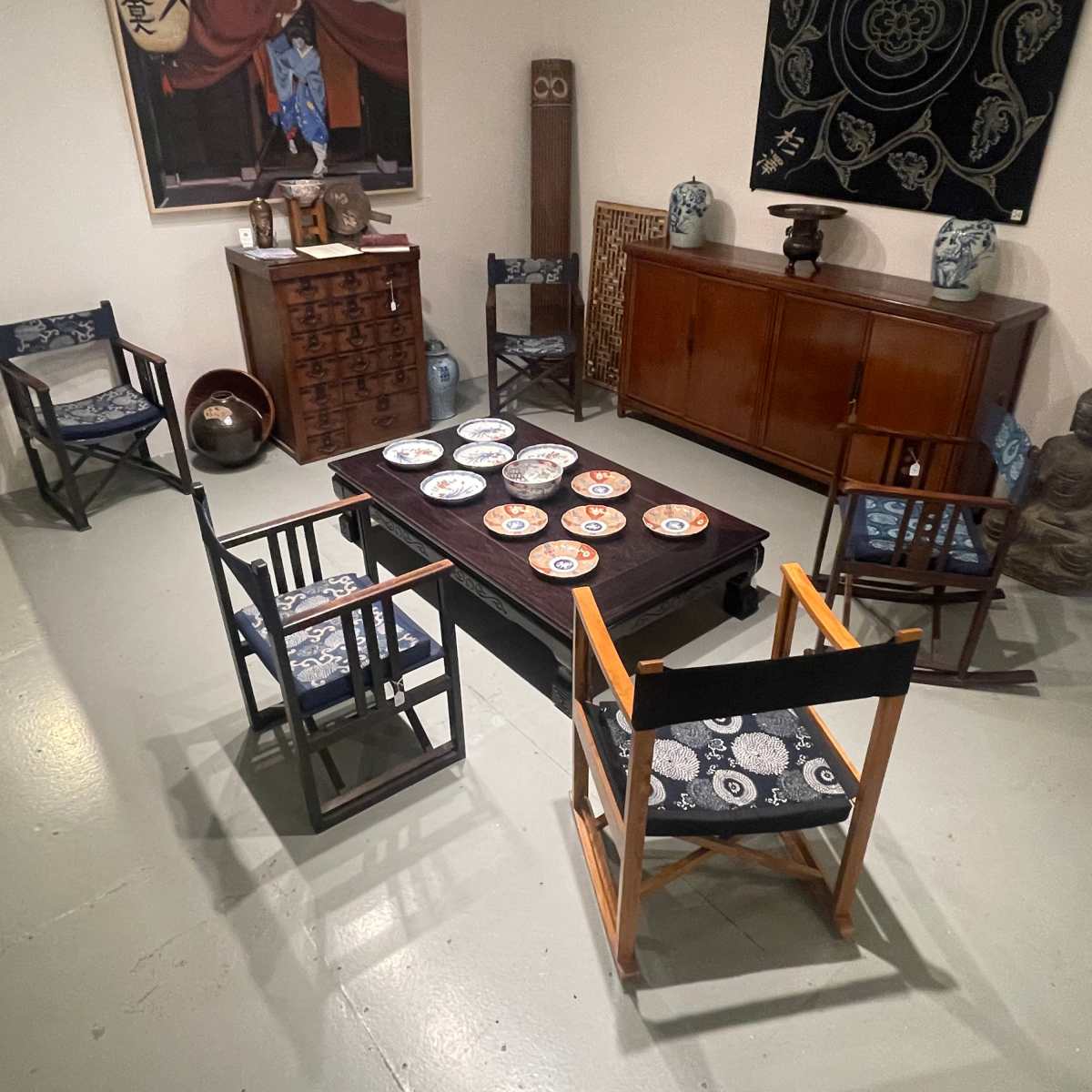 A room furnished with a wooden table surrounded by four chairs, decorative plates arranged on the table, and various pieces of artwork and decor in the background.