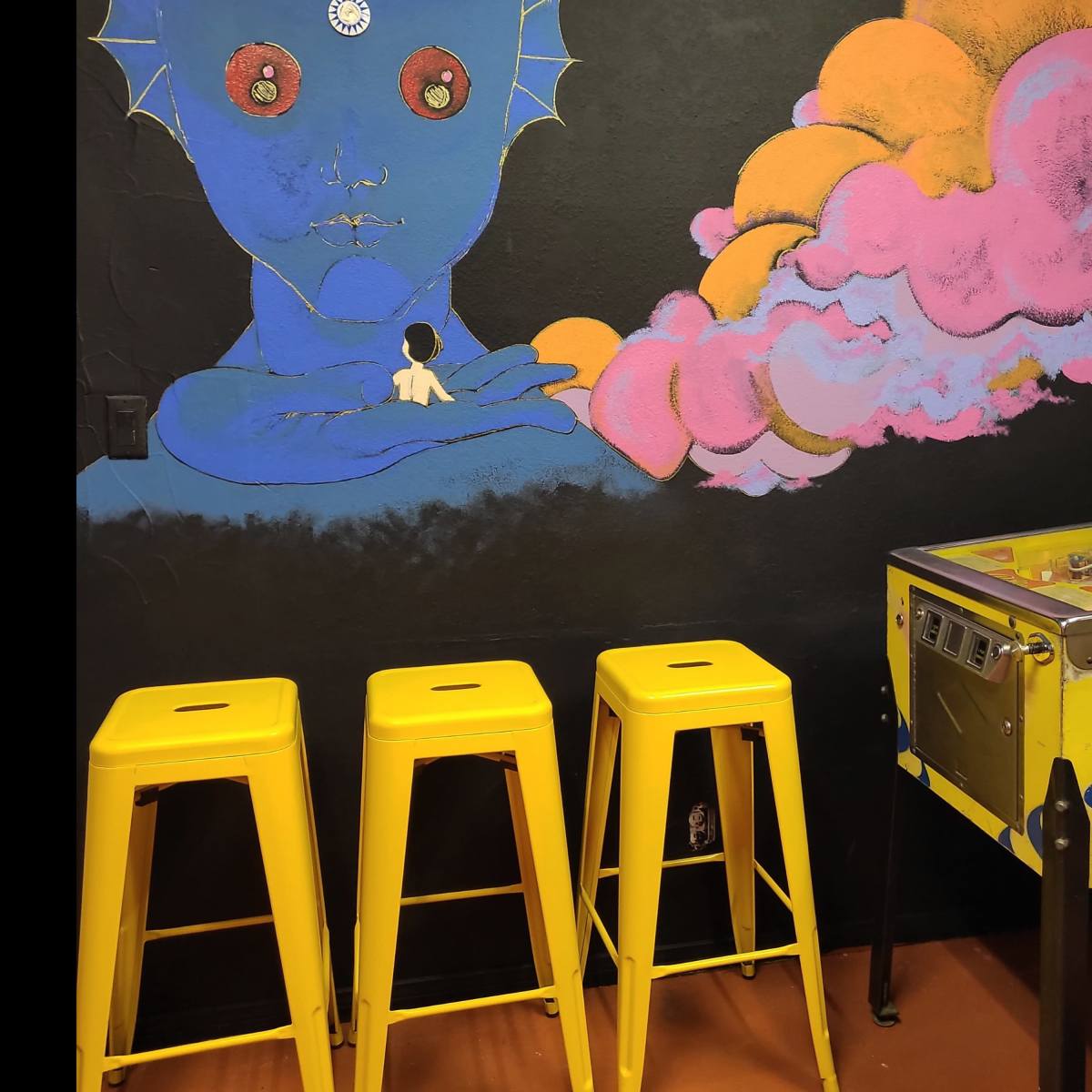 A mural of a blue-skinned figure with a cosmic head is painted on a black wall beside three yellow stools and a pinball machine.