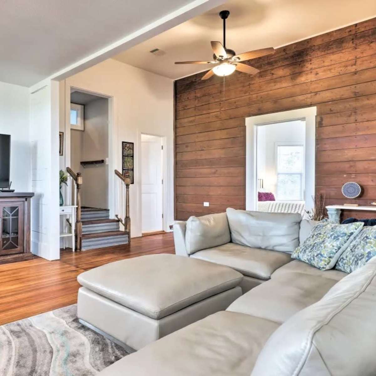 The living room features a sectional sofa, a flat-screen TV on a wooden stand, and a staircase leading to an upper level, with a mirror reflecting the space.
