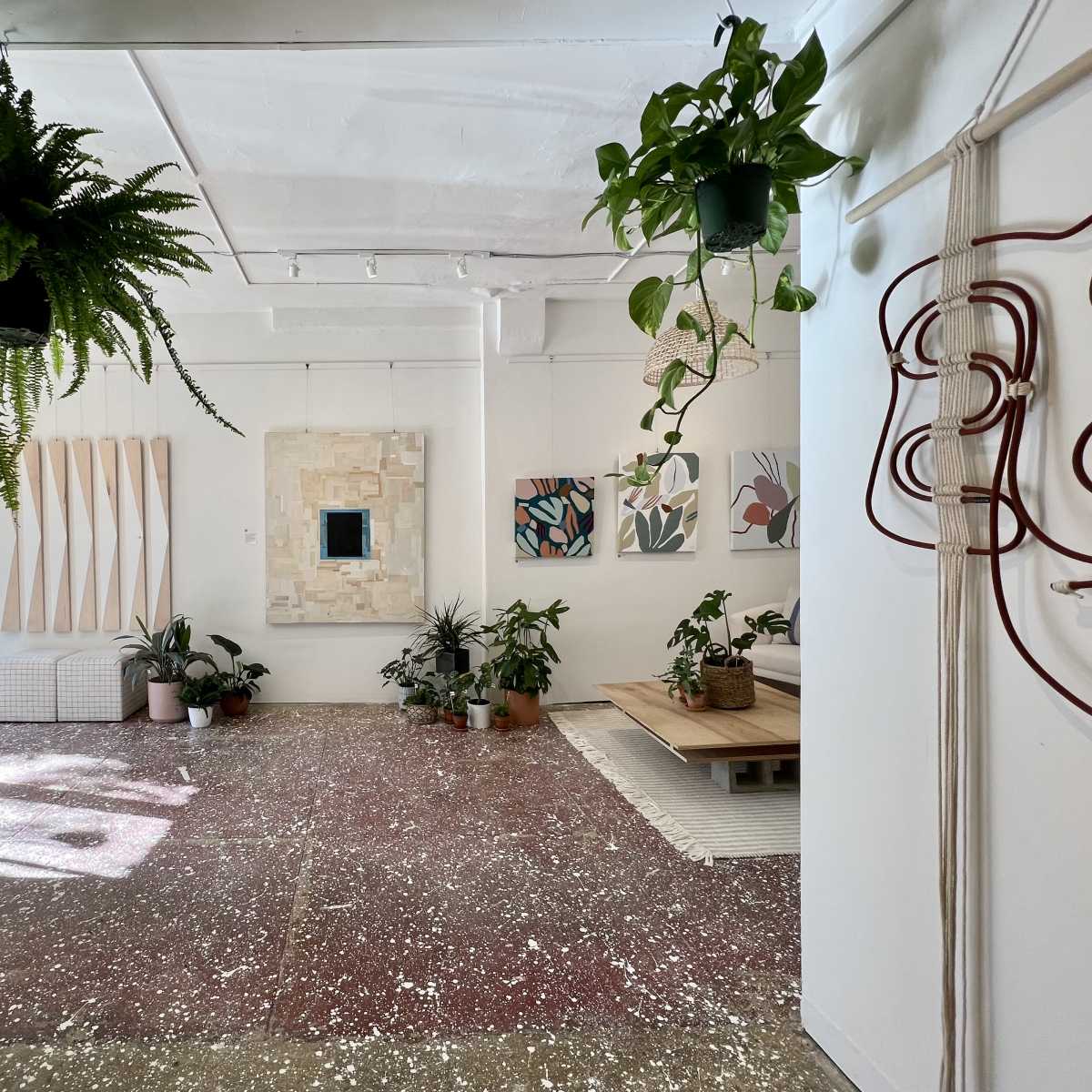 A brightly lit contemporary interior space featuring plants, artwork on the walls, and a mixture of furniture and decorative elements on the floor.