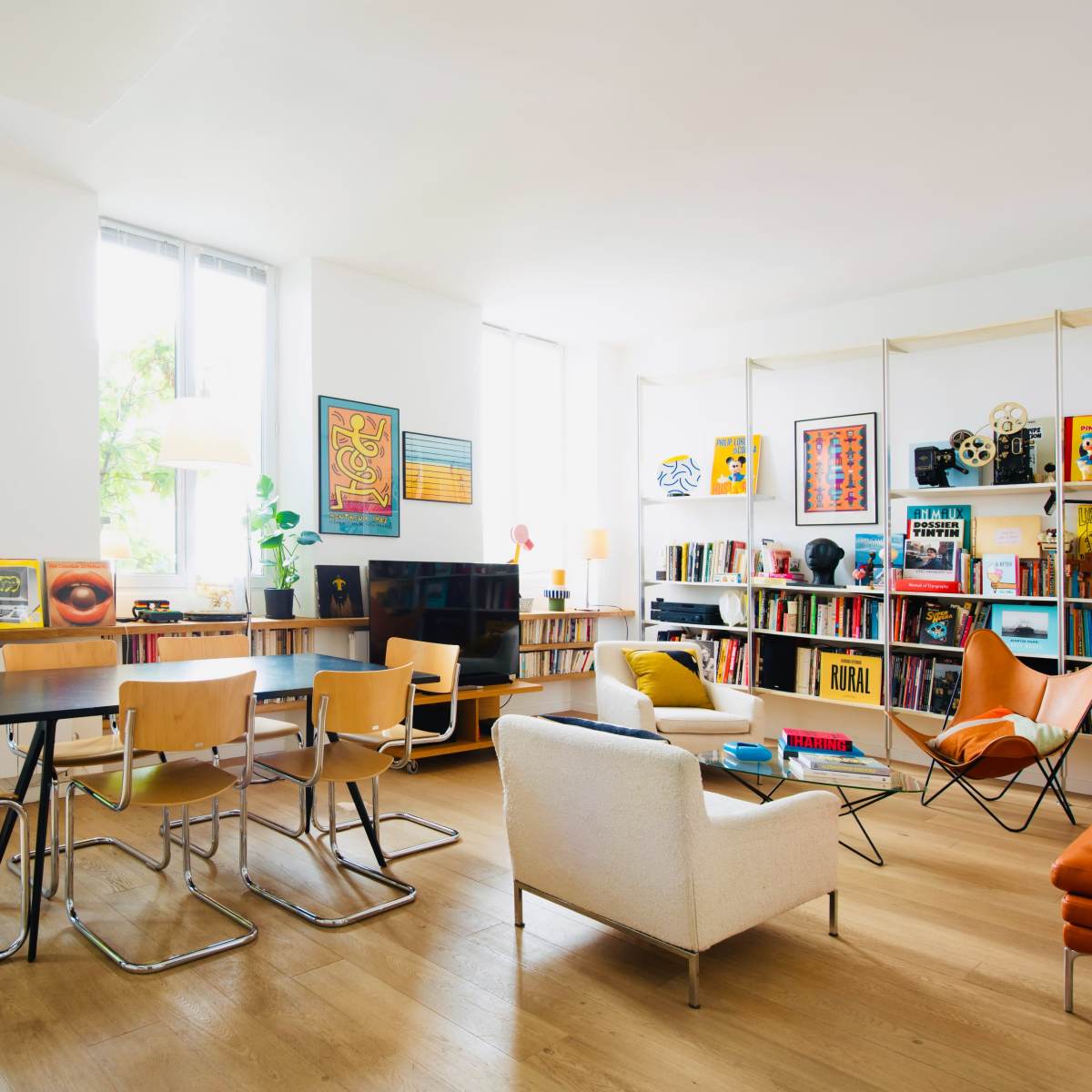 A bright living room features a dining table with four chairs, a comfortable armchair, and a bookshelf filled with various books and art pieces.