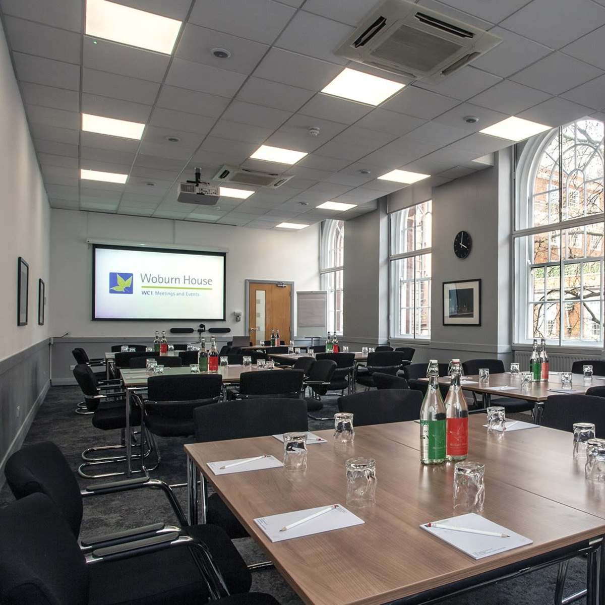 A conference room set up with several tables arranged for a meeting, featuring chairs and water bottles on each table, with a screen displaying "Woburn House" in the background.
