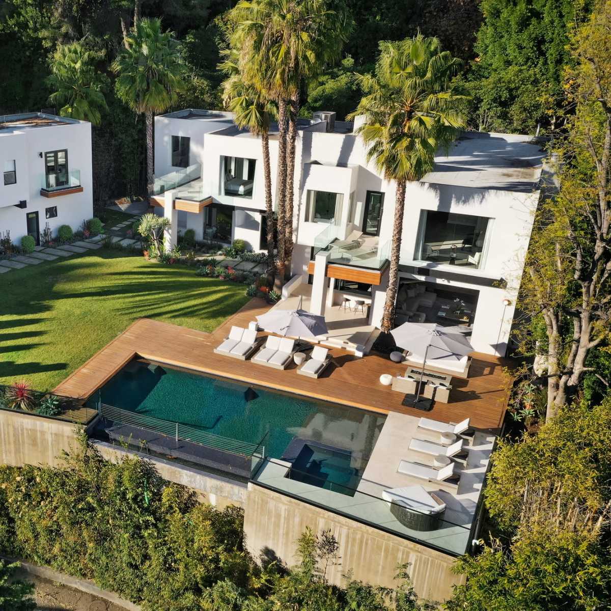 A modern two-story house with a grassy lawn, palm trees, and a swimming pool surrounded by a wooden deck.