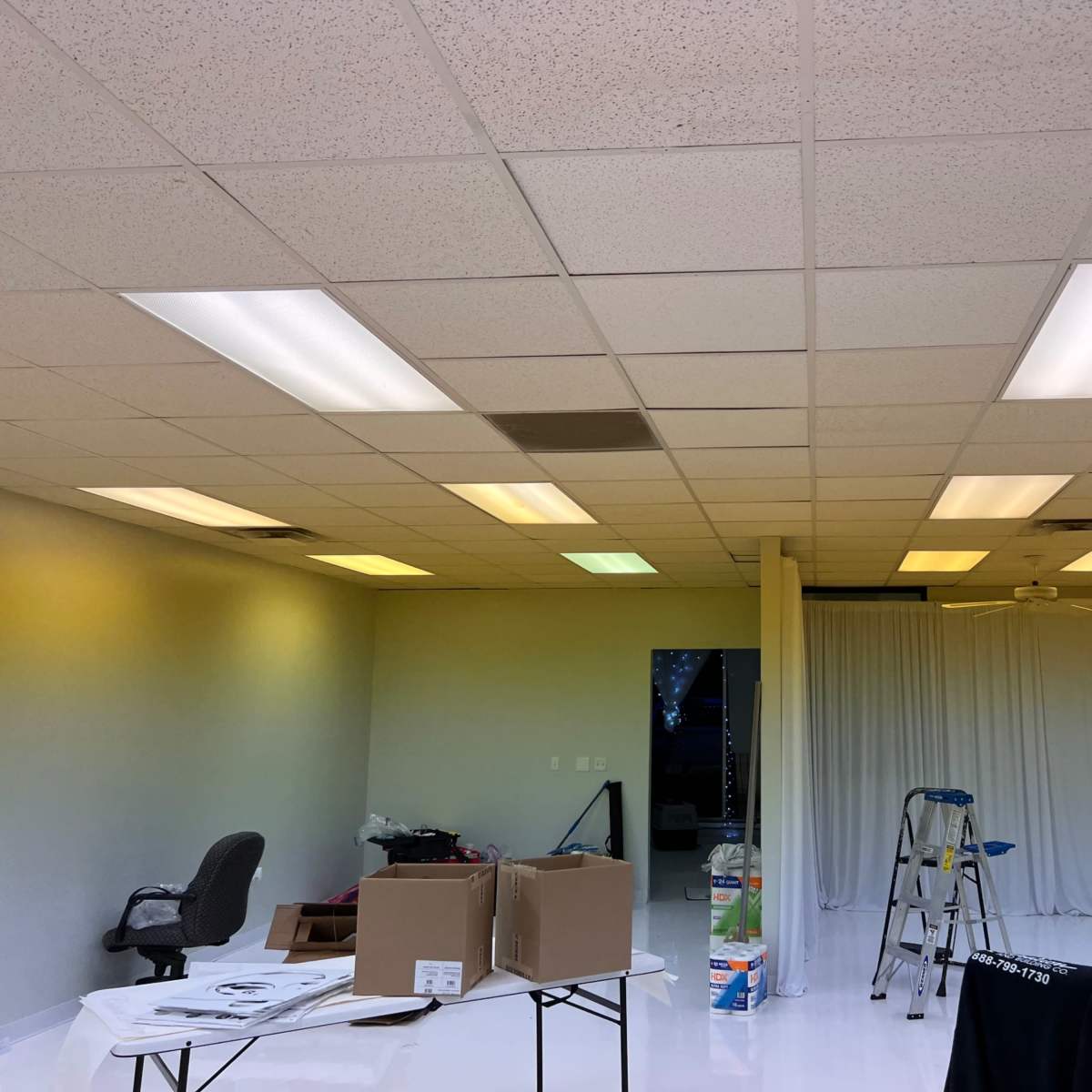 An empty room with fluorescent lighting, a ladder, some cardboard boxes, and a partially draped white curtain.