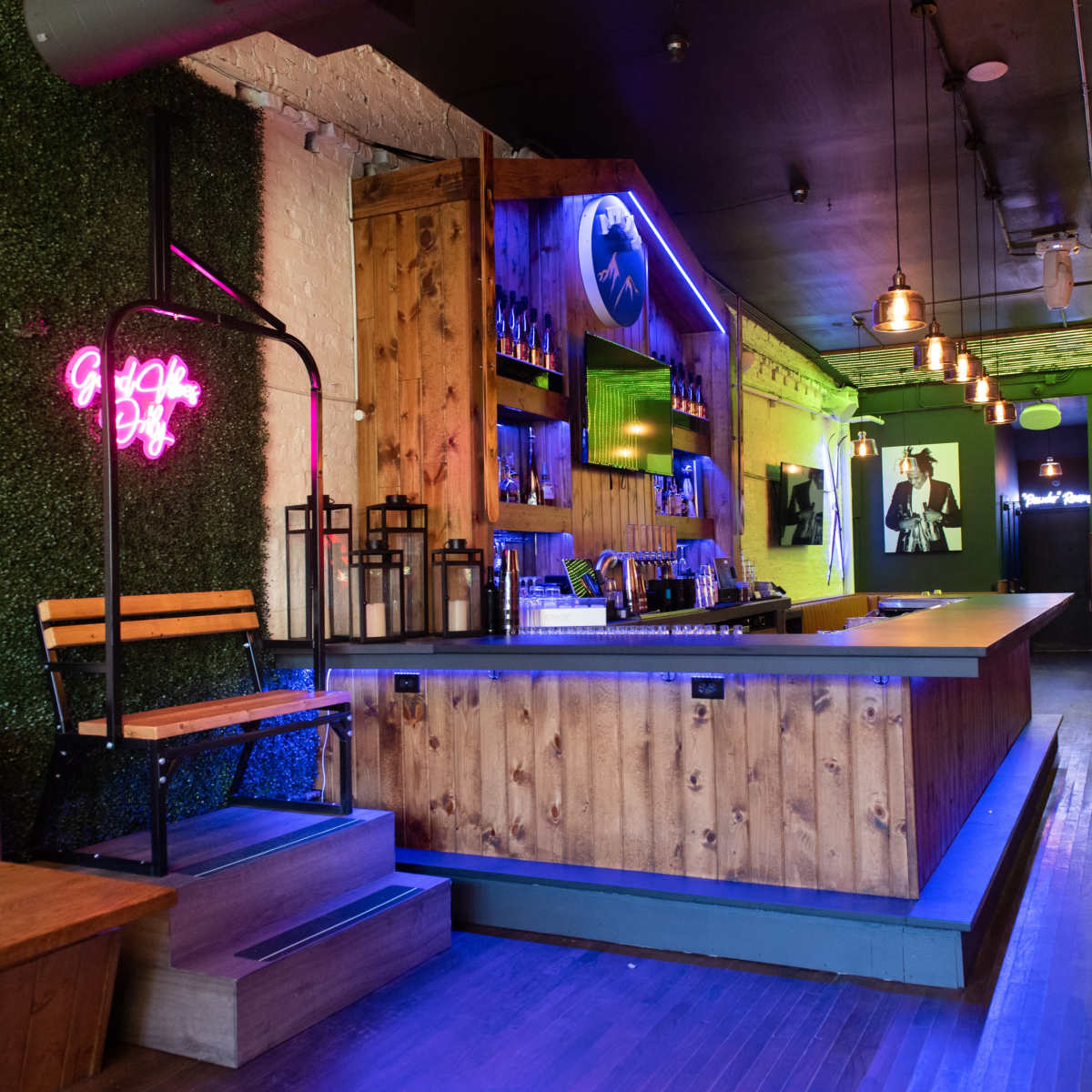 A stylish bar area featuring a wooden counter, neon signs, and a combination of green wall foliage and colorful lighting.