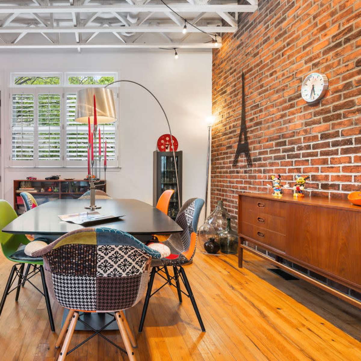 A modern dining area with a large table surrounded by colorful chairs, hardwood floors, and exposed brick walls.