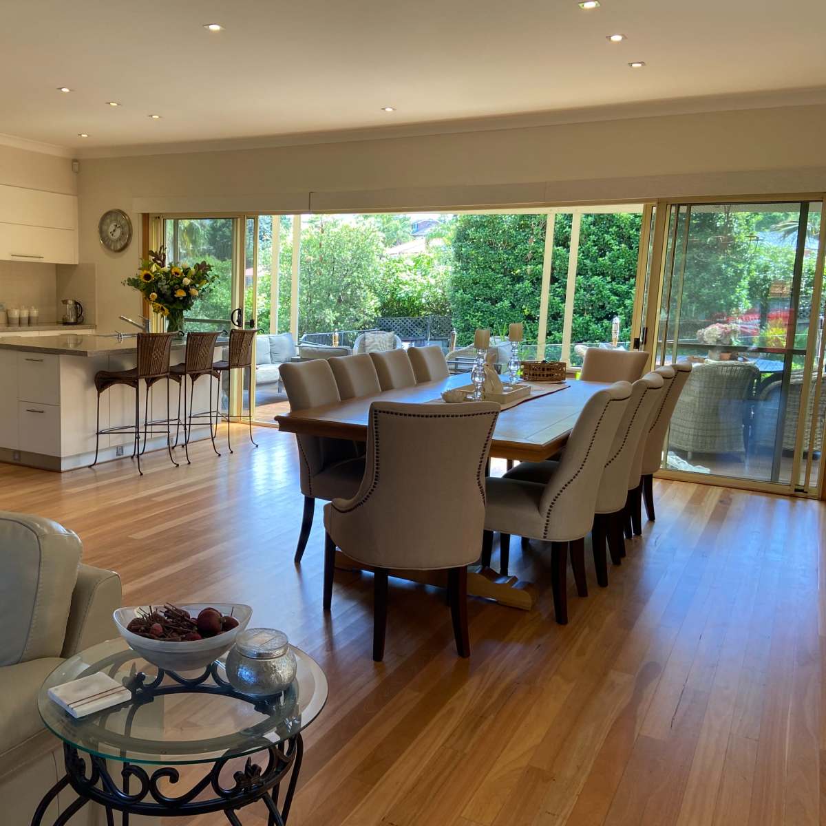 A modern, open-plan living and dining area with a kitchen, wooden floors, a large dining table, and sliding glass doors leading to an outdoor space.