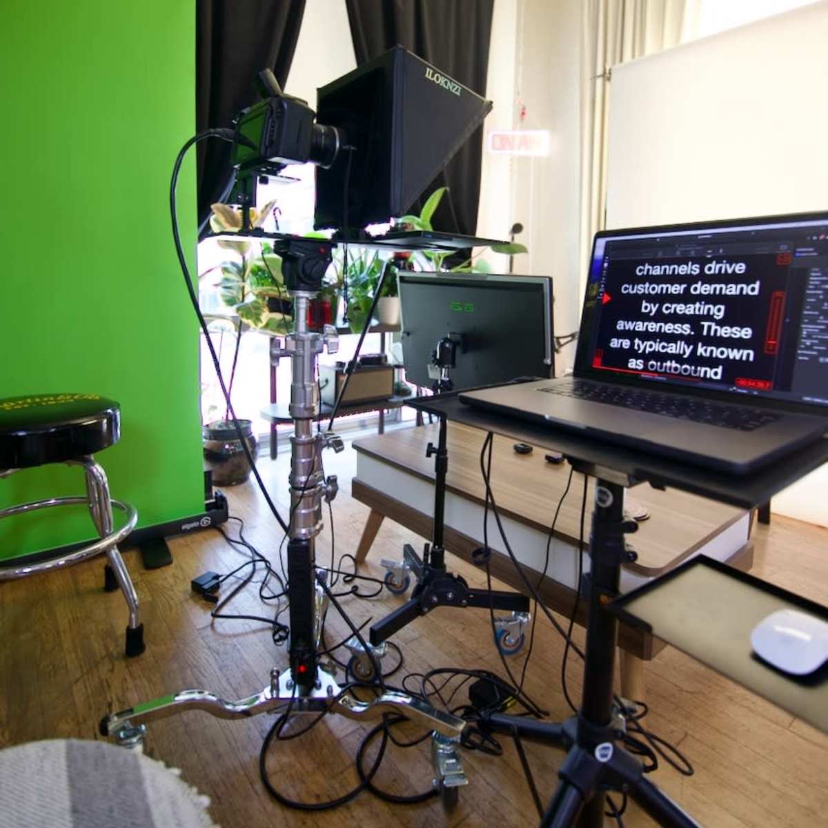 A video production setup is arranged with a green screen, a monitor displaying text, and an adjustable camera on a tripod, surrounded by plants and basic furnishings.
