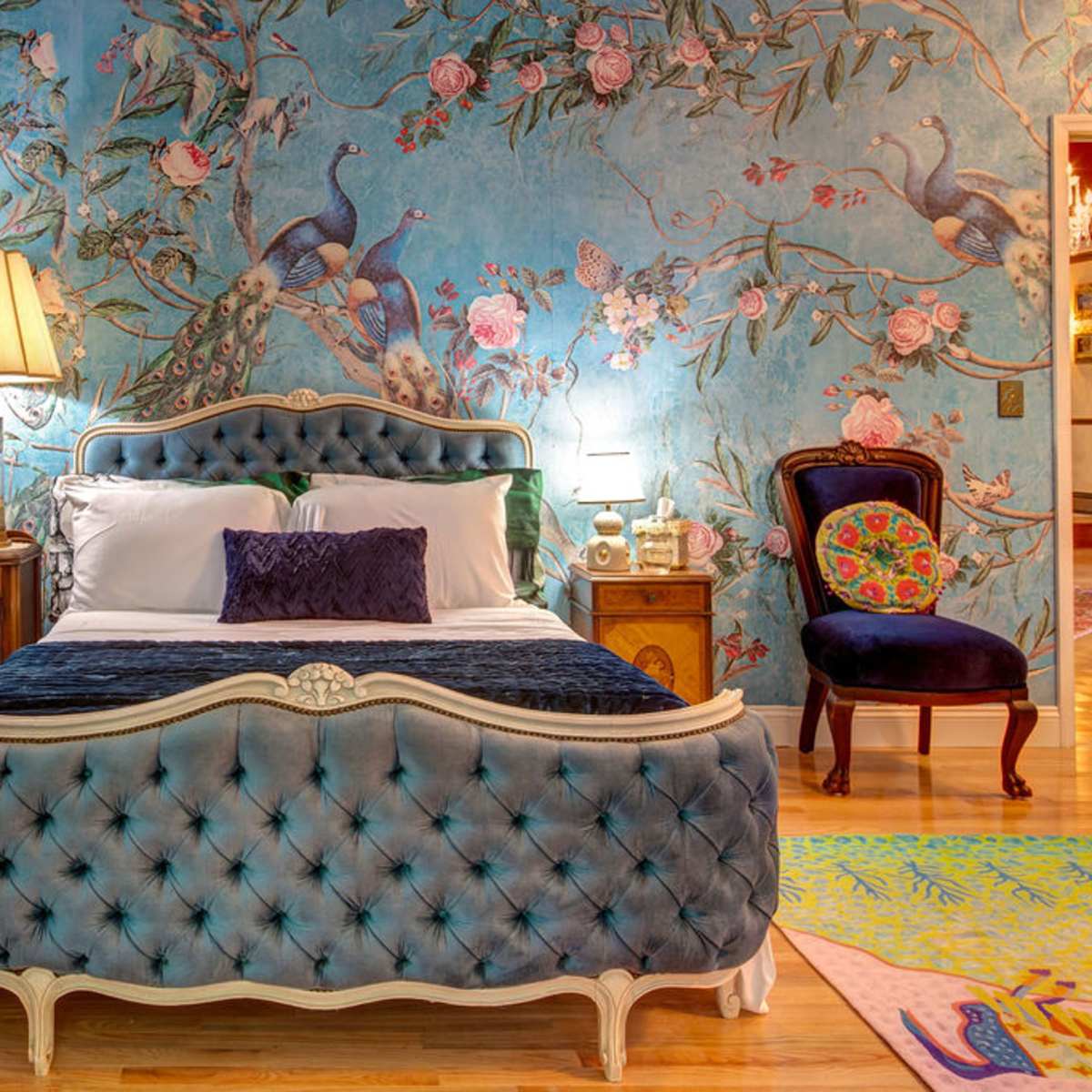 A bedroom featuring a blue tufted bed with a decorative headboard, a floral wallpaper backdrop, and a combination of luxurious furniture elements.