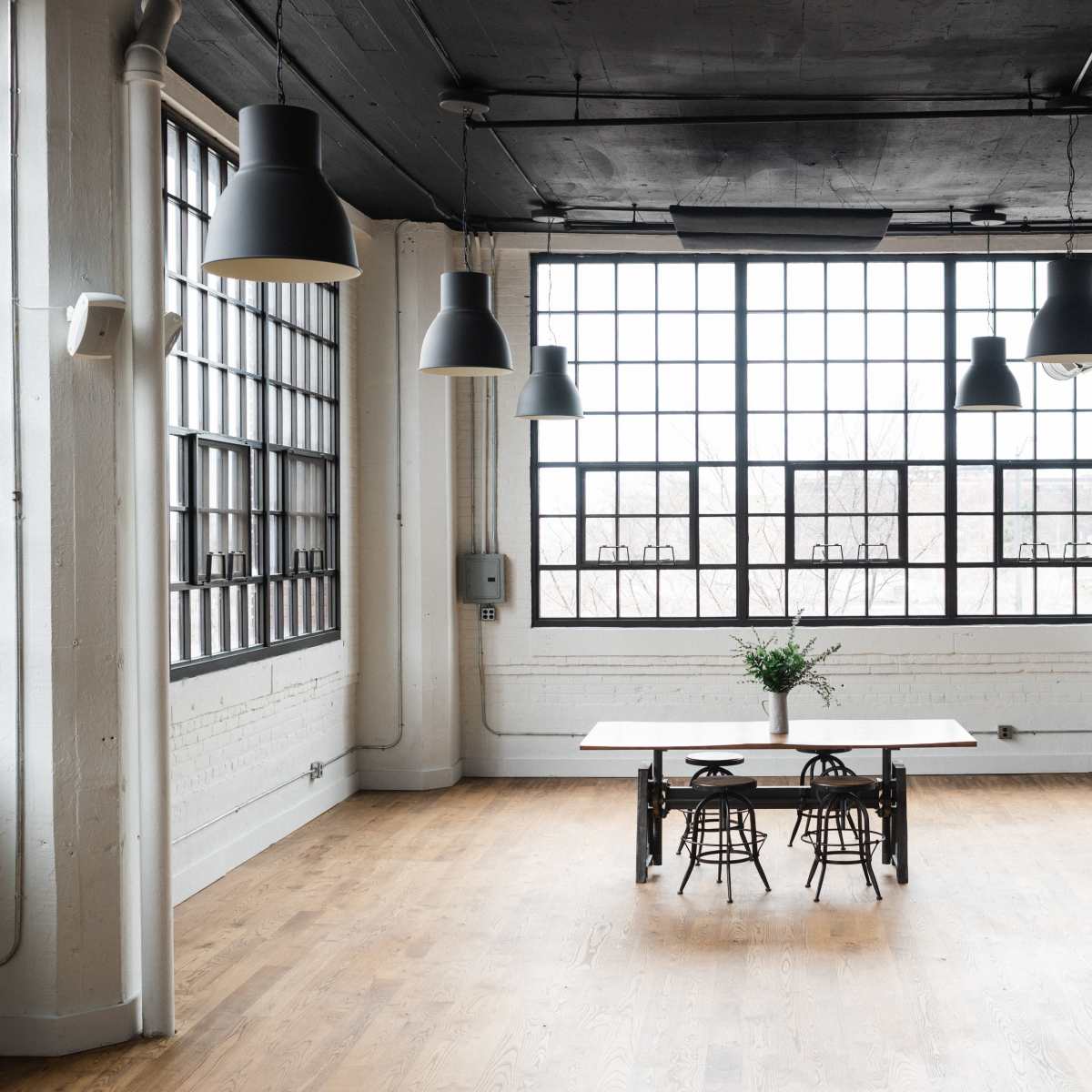 A spacious, bright room features a central wooden table surrounded by metal chairs, with large windows lining the walls.