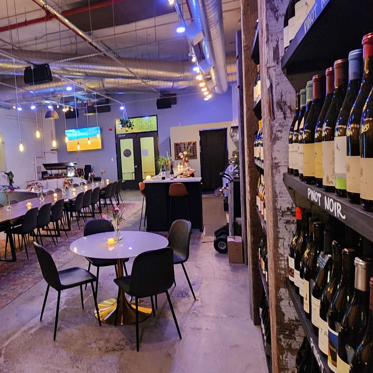 A modern wine bar with a long table and chairs in the center, flanked by shelves of wine bottles on one side and a bar area on the other.