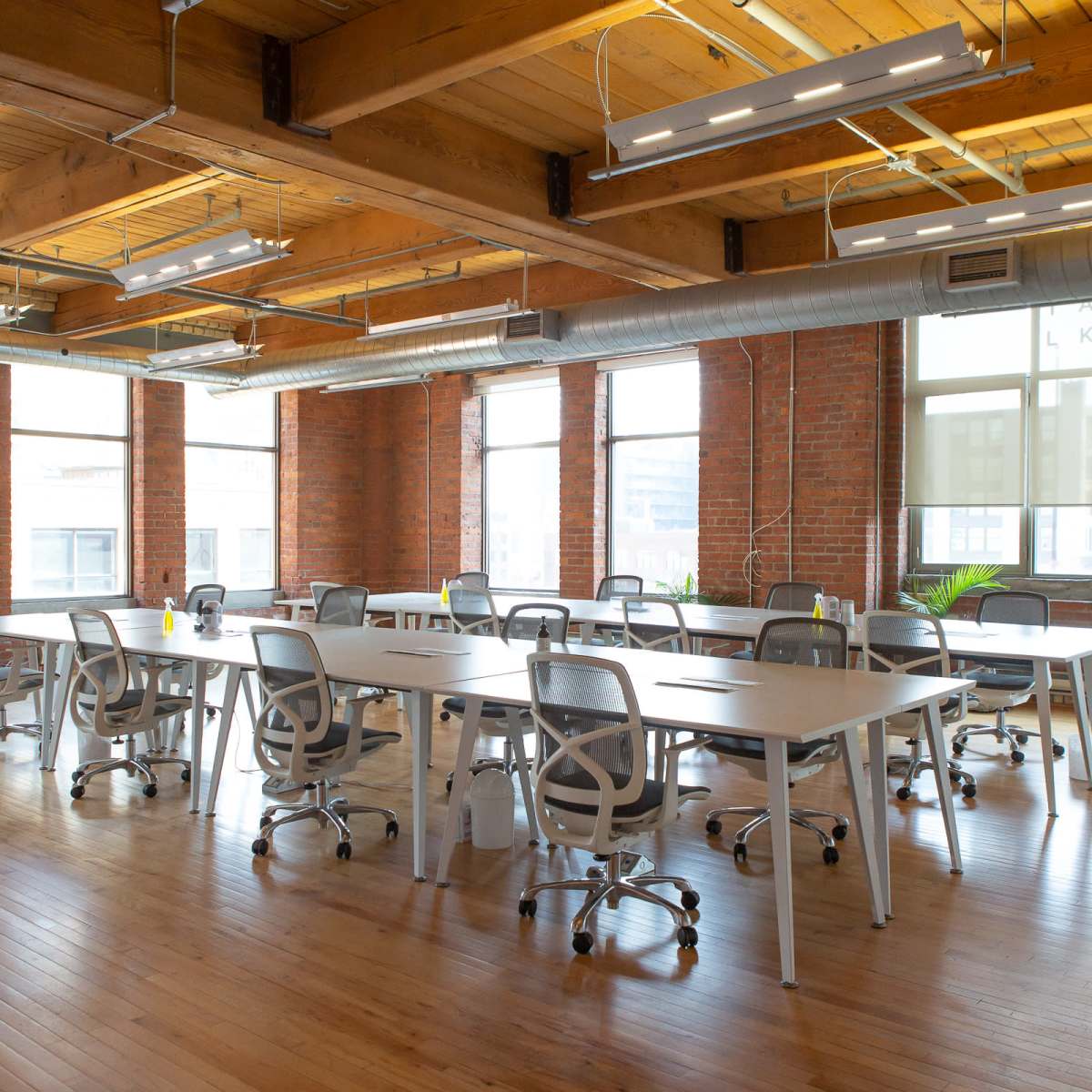A spacious office with wooden floors and large windows, featuring multiple work tables and ergonomic chairs arranged in rows.