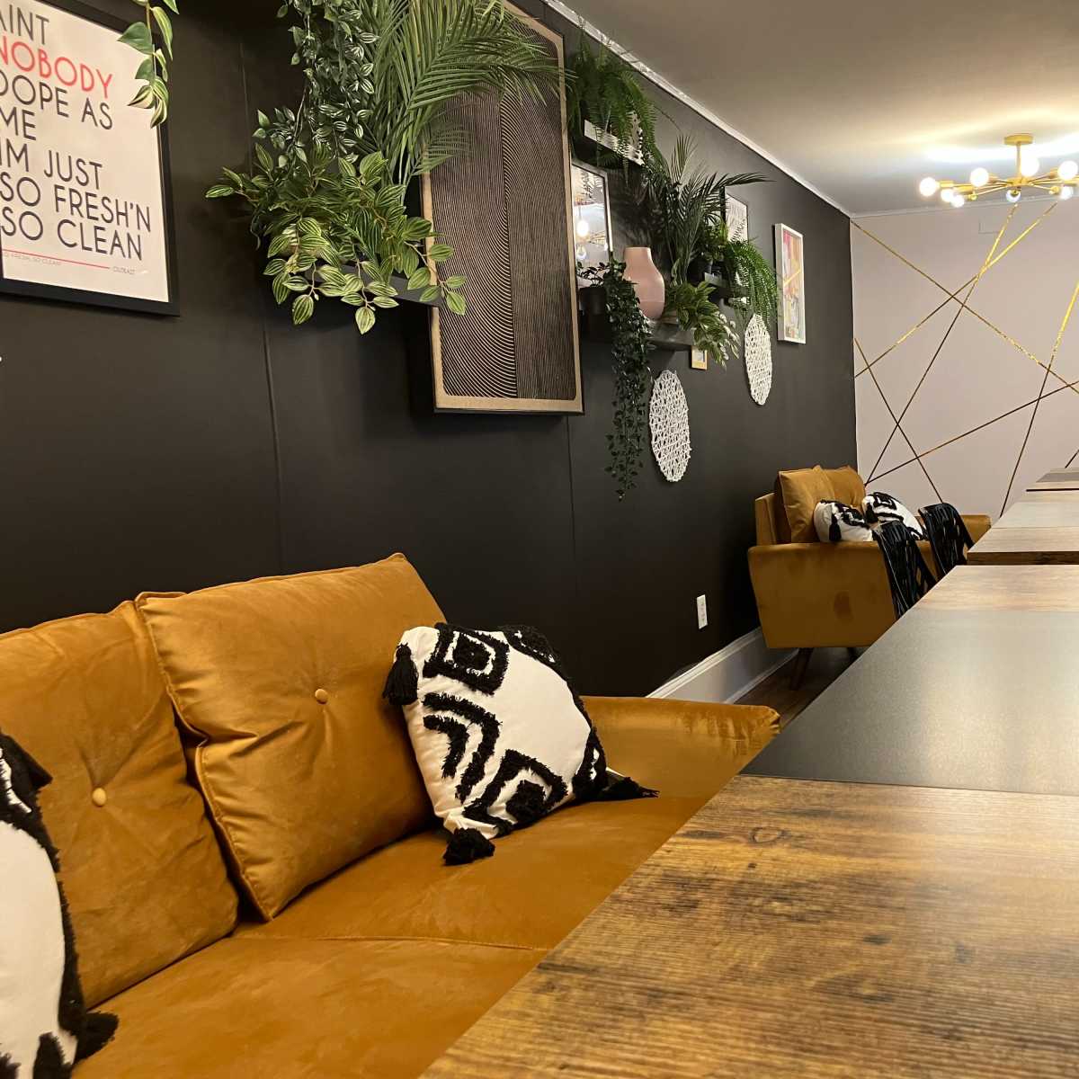A long conference table is set in a modern meeting room, adorned with plants and decorative art, while brown sofas with patterned cushions provide seating along one side.