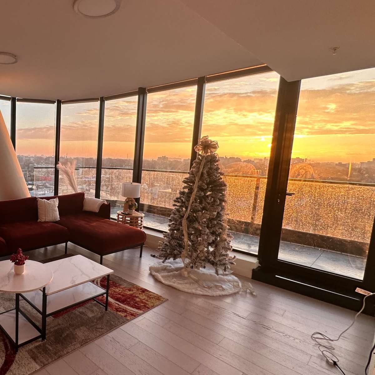 A modern living room with large windows overlooking a sunset, a red sofa, a white coffee table, and a Christmas tree decorated with lights.
