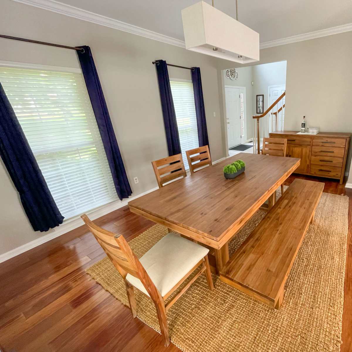 A well-furnished dining area featuring a large wooden table with benches, accompanied by a single chair, set against a backdrop of windows with white blinds and dark curtains.