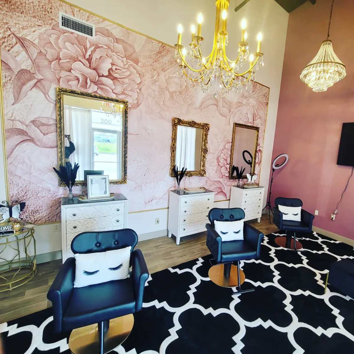 A modern beauty salon featuring three black chairs, ornate mirrors, and a floral-themed accent wall, all illuminated by chandeliers.