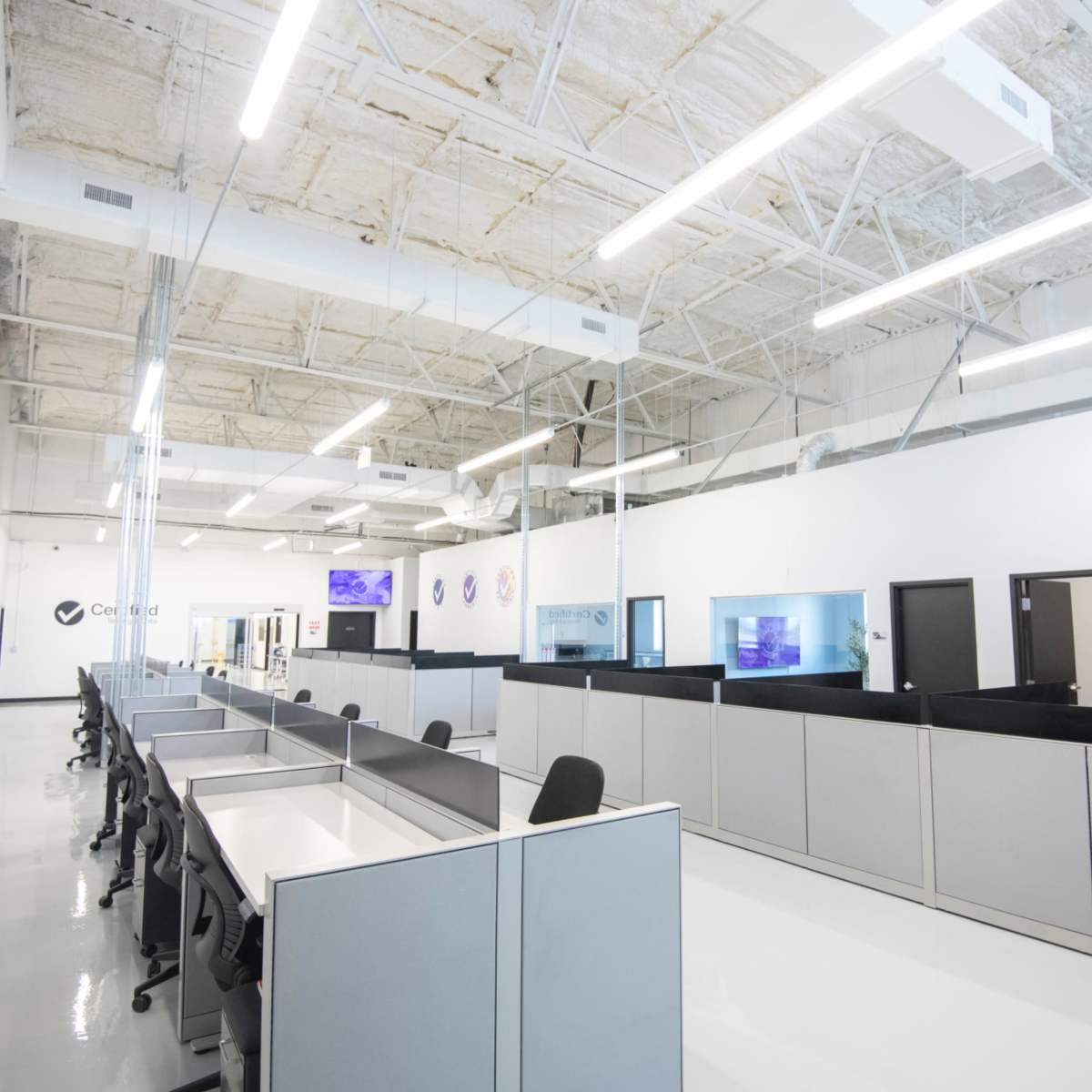 A spacious, modern office layout with cubicles and high ceilings, featuring white walls and bright overhead lighting.
