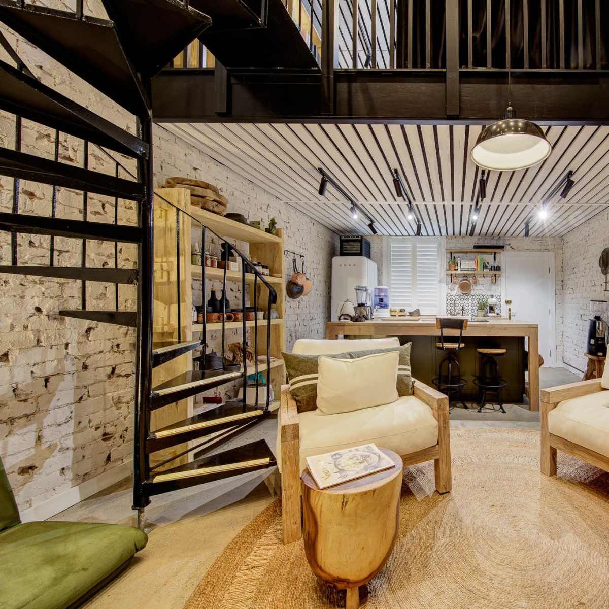 A modern interior space with a spiral staircase, two wooden chairs, and a kitchen area, featuring exposed brick walls and a round rug.