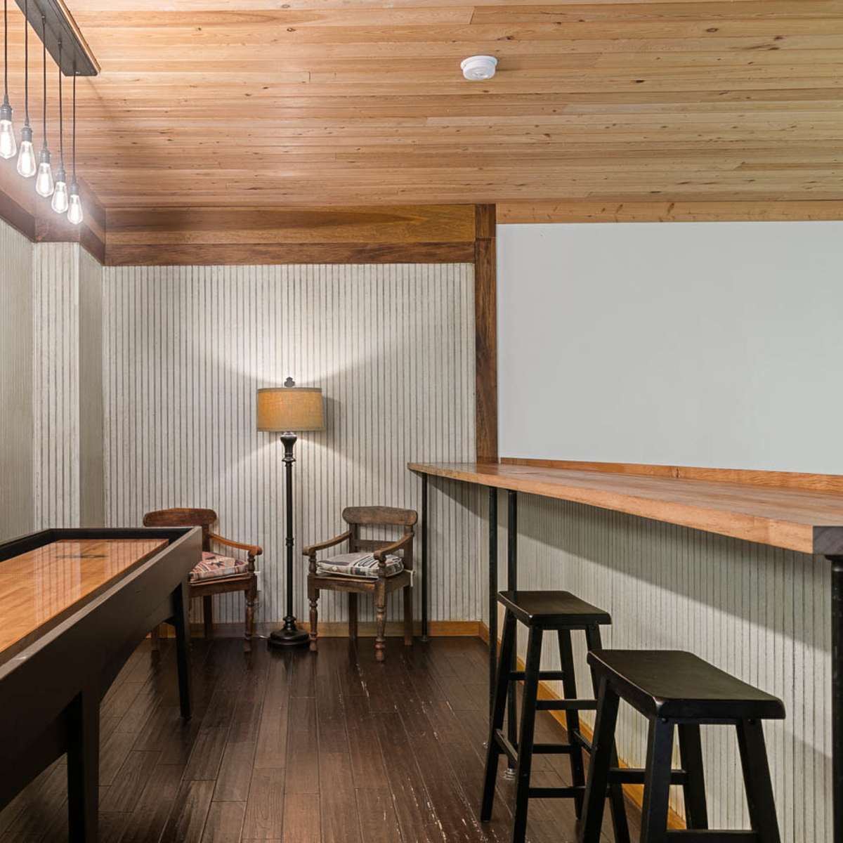 A neatly designed room featuring a wooden bar top, two black stools, a lamp, and two chairs, all situated under a ceiling with exposed bulbs and a stained glass window.
