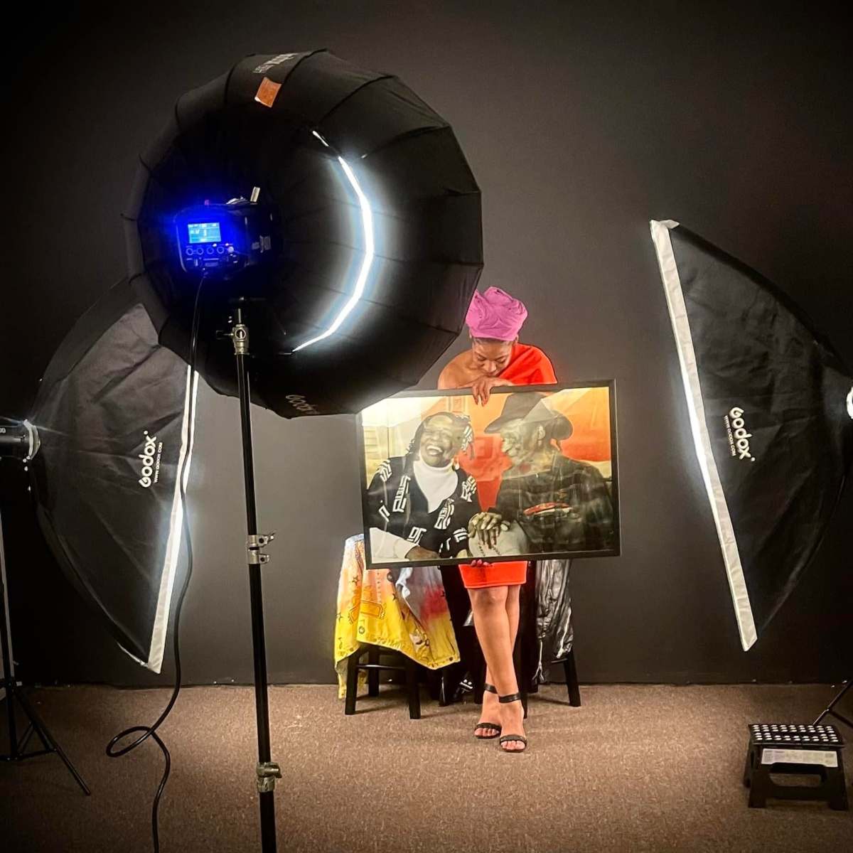A person holds a framed painting in a photography studio, surrounded by softbox lights and a dark backdrop with motivational text.