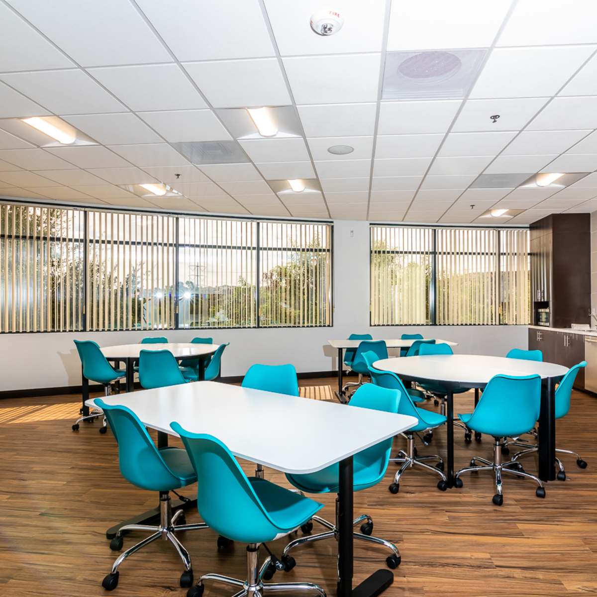 A modern dining area with multiple white tables and turquoise chairs, lit by natural light through large windows.