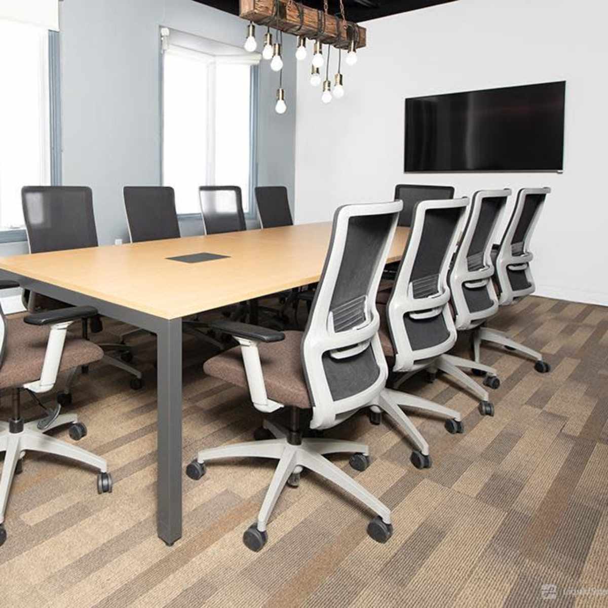 A long conference table with rolling chairs is set up in a well-lit meeting room featuring a television and windows.