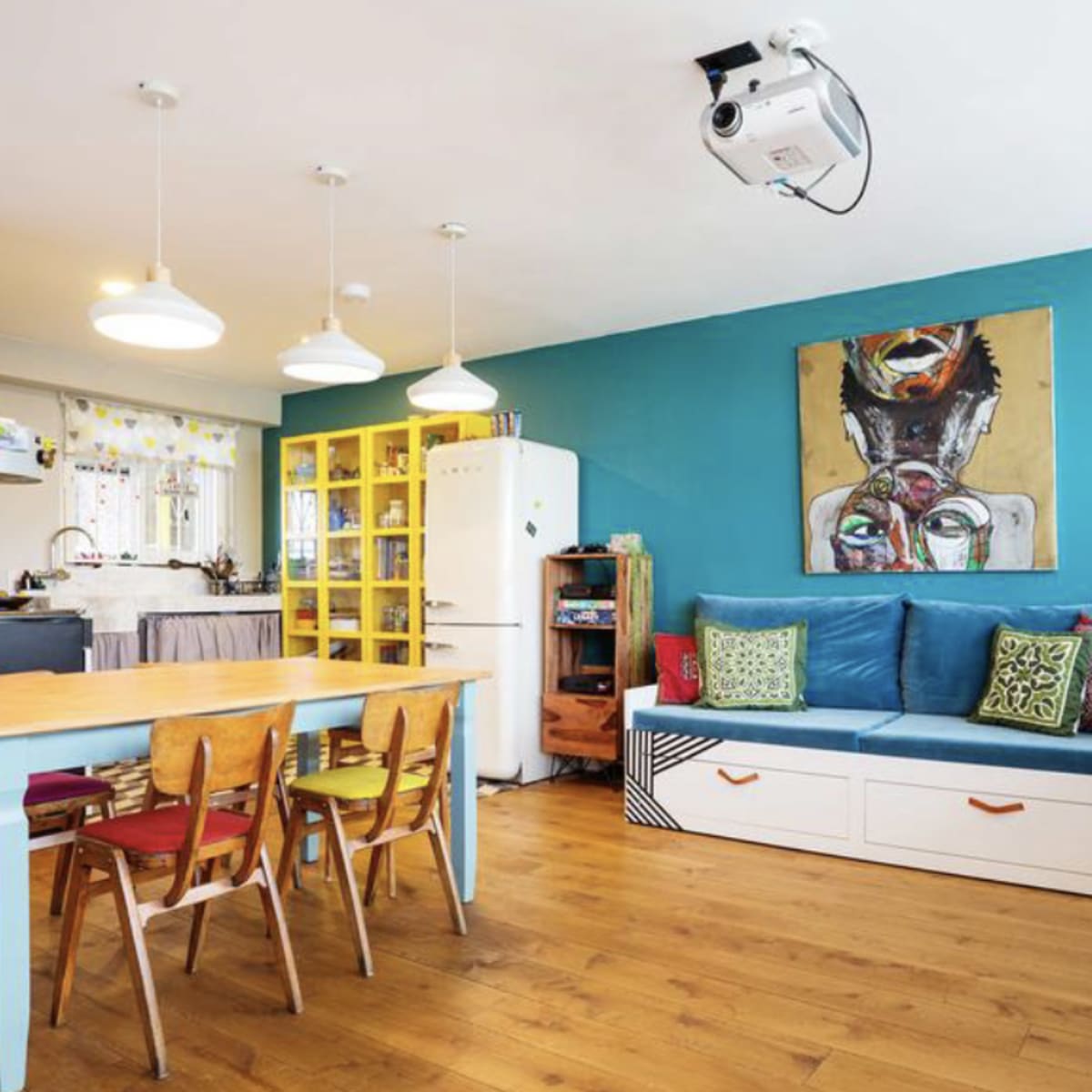 A brightly colored living space featuring a dining table with chairs, a blue accent wall, a projector mounted on the ceiling, and various decorative elements.