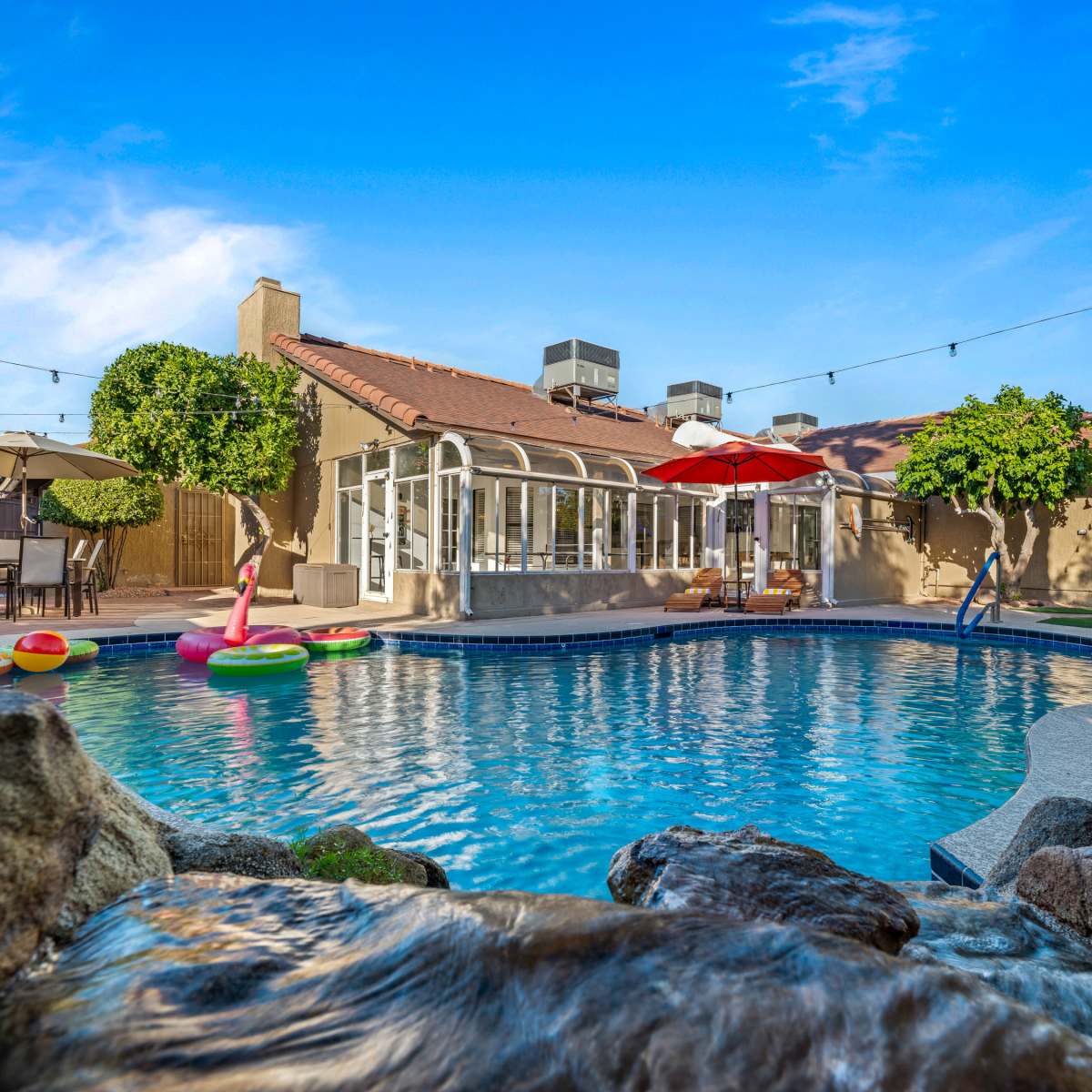A clear pool with inflatable floats is situated in a backyard alongside a house with a patio and umbrellas.