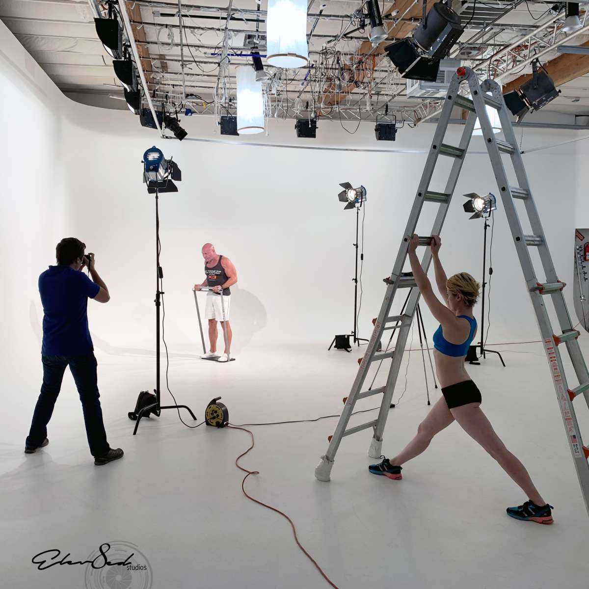 A male model is posing with lighting equipment set up in a photography studio while a female photographer on a ladder adjusts the shot.