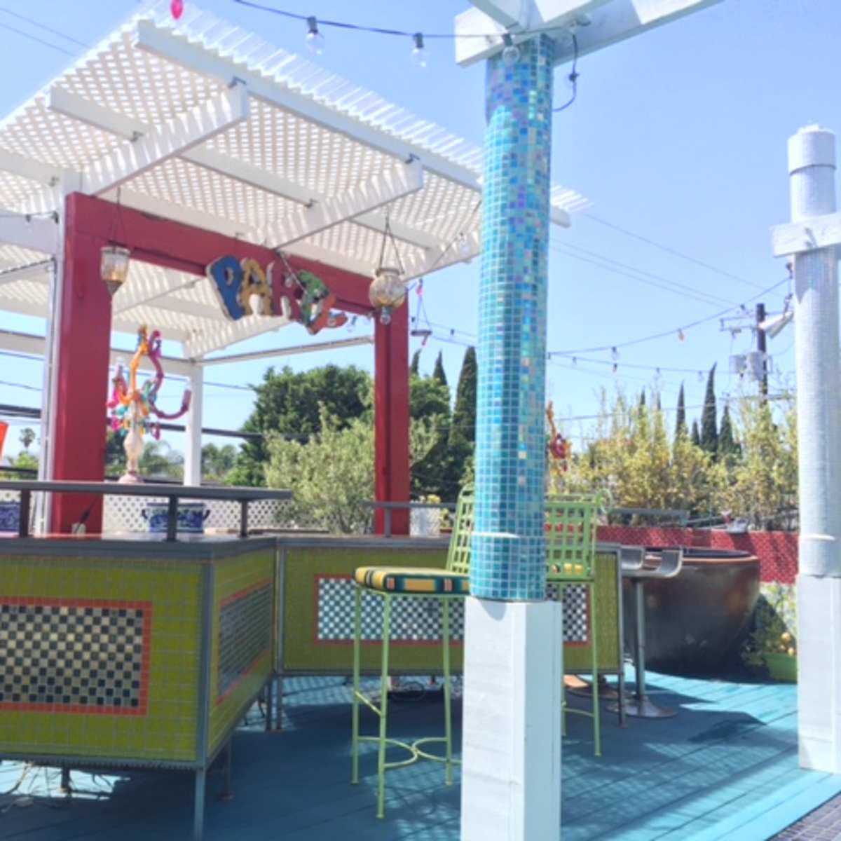 A colorful outdoor bar area features a large shaded pergola with decorative elements, surrounded by greenery and bright furniture.