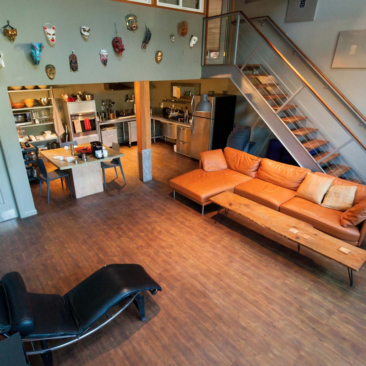 A spacious, open-concept living area with a brown leather sofa, a wooden coffee table, a dining area, an industrial kitchen, and a staircase leading to a loft.
