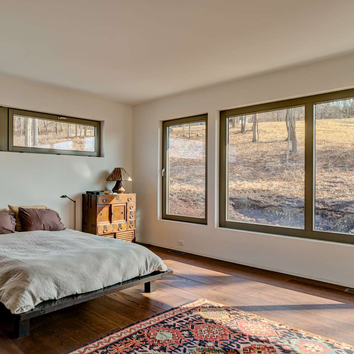 A bedroom features a low bed with brown bedding, a wooden dresser, and large windows overlooking a grassy landscape.