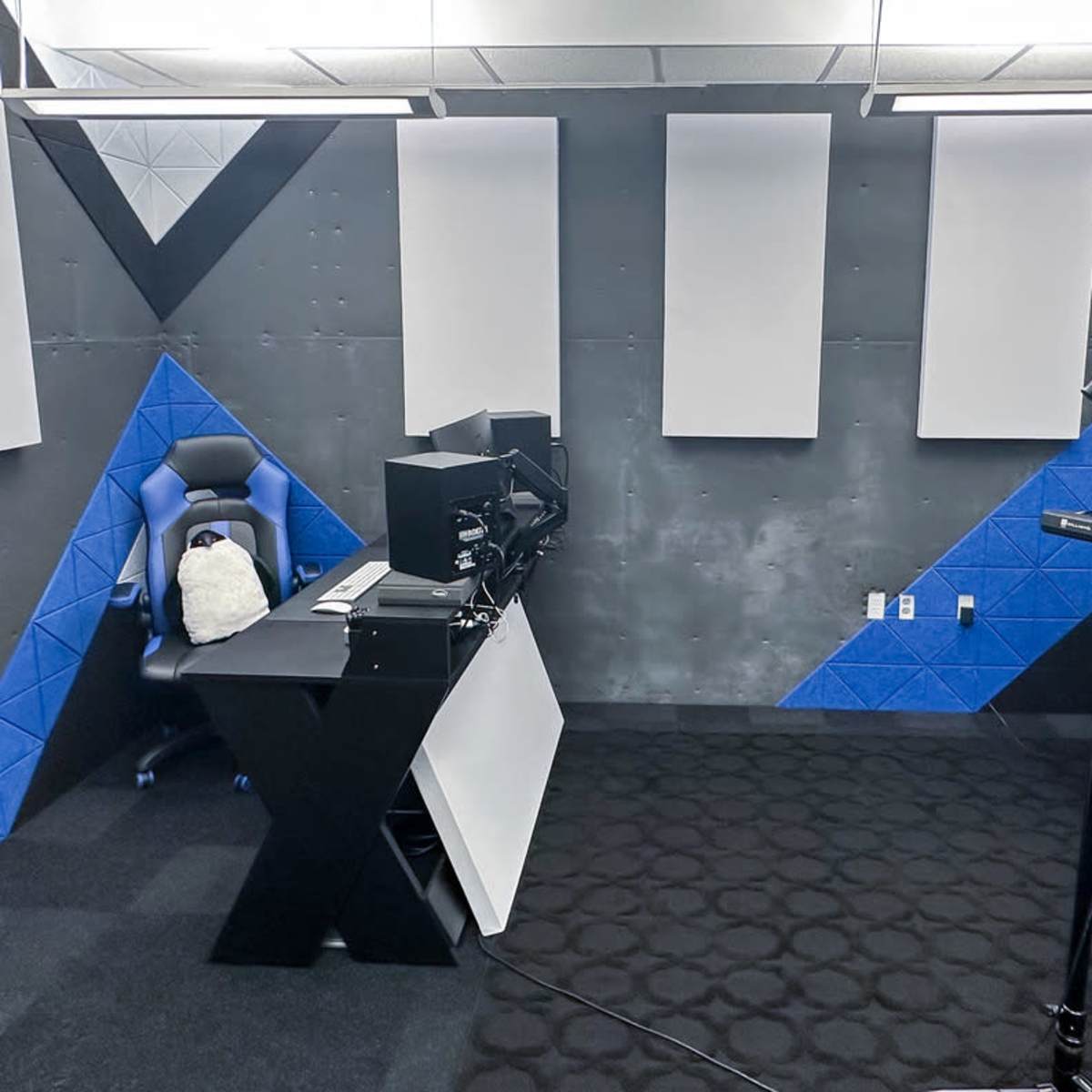 A stylish recording studio with a blue and black color scheme, featuring a microphone on a stand, a gaming chair, and soundproofing panels on the walls.
