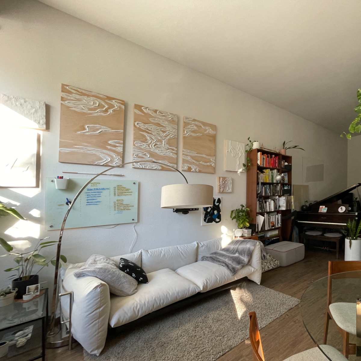 A cozy living room with a white sofa, bookshelf, piano, and various plants arranged around the space.
