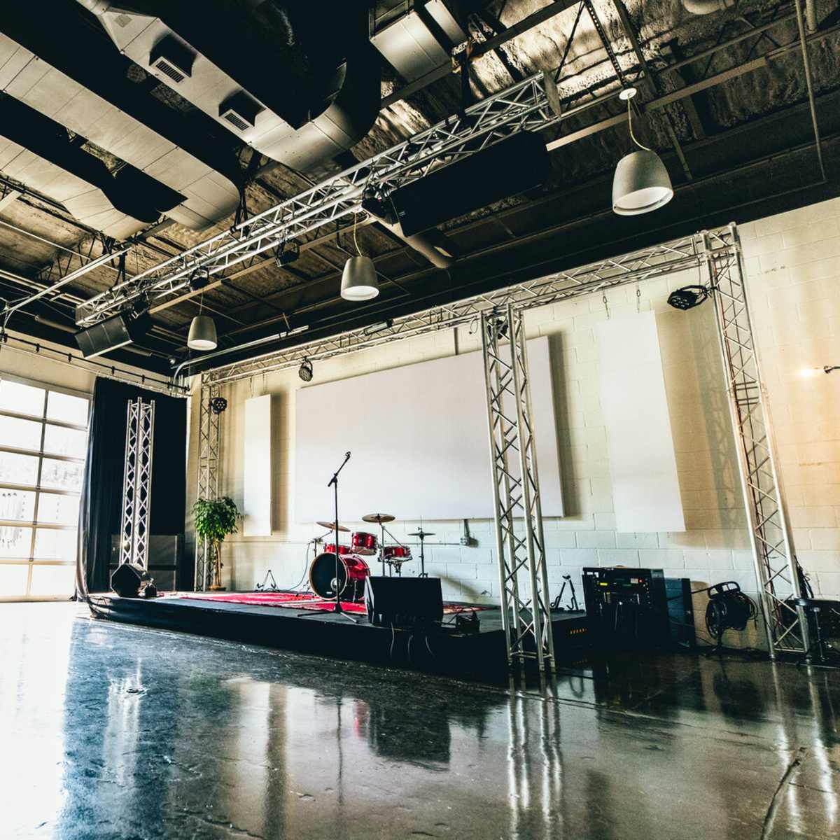 A spacious performance space featuring a stage with a drum set, black curtains, and a large window allowing natural light to enter.