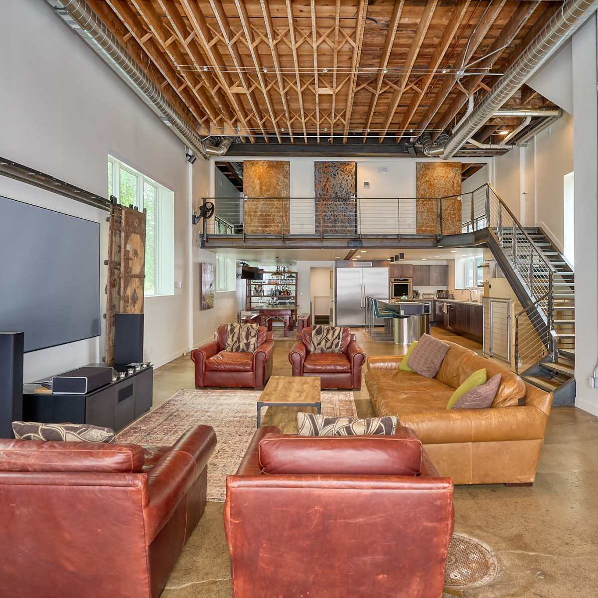 The spacious living area features leather sofas, a large rug, and an open staircase with a kitchen visible in the background.