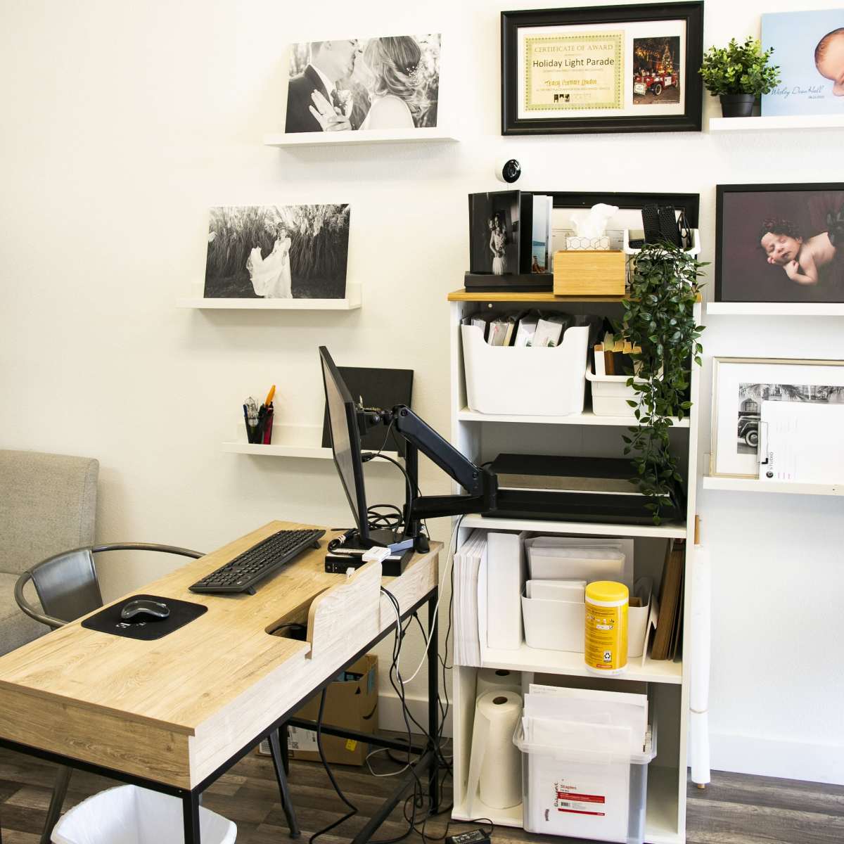 A small office workspace featuring a wooden desk with a computer, shelves displaying photographs and a certificate, and storage units with various supplies.