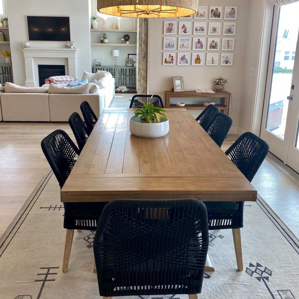 A wooden dining table surrounded by black chairs is centered in a well-lit room with a large pendant light, a wall of framed photos, and a cozy seating area in the background.