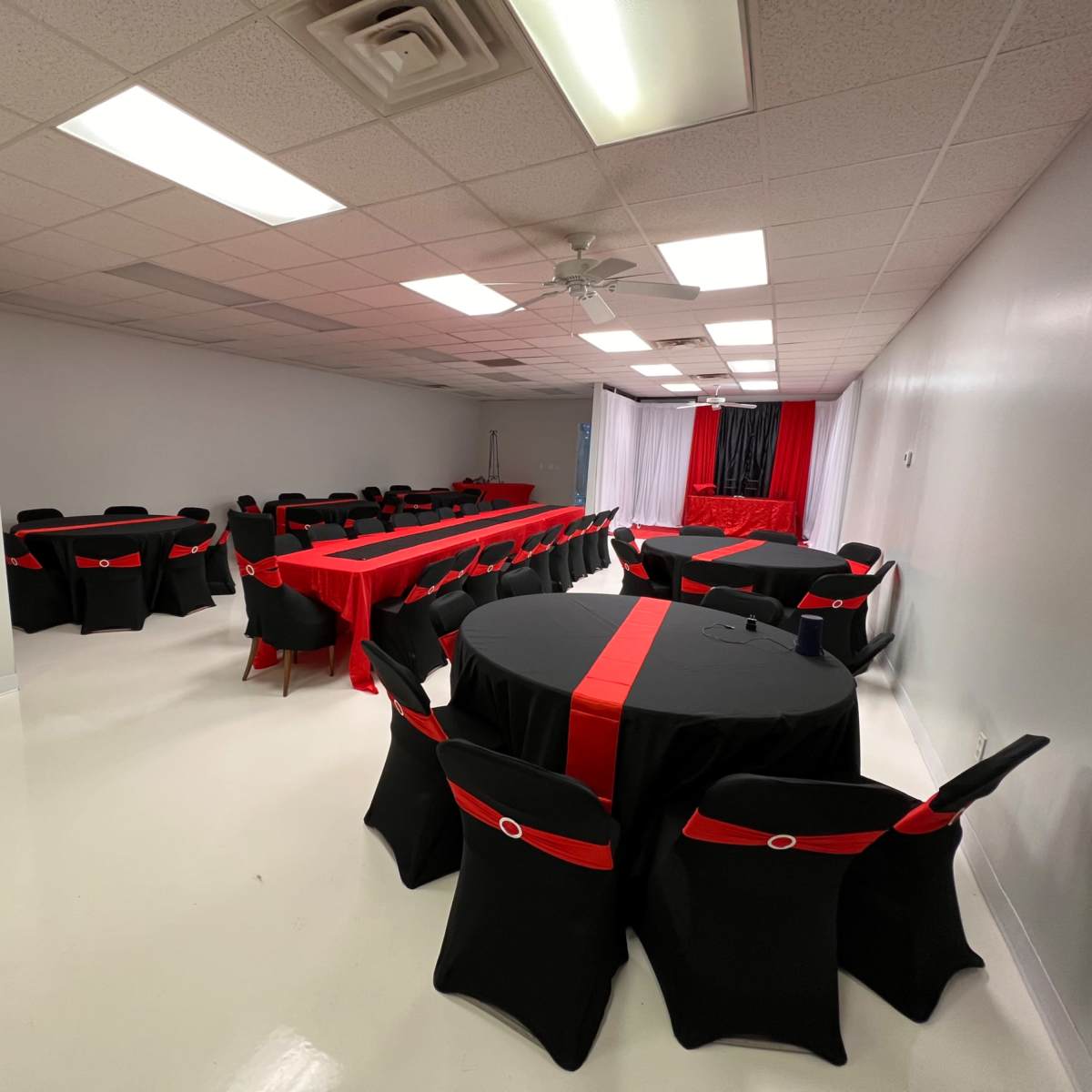 A banquet hall set up with round tables covered in black tablecloths adorned with red ribbons, illuminated by overhead fluorescent lights.