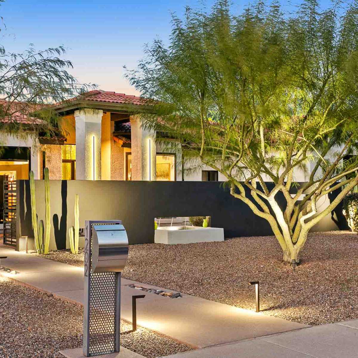 A modern single-story home surrounded by desert landscaping, featuring a pathway lined with lights and a mailbox near the entrance.