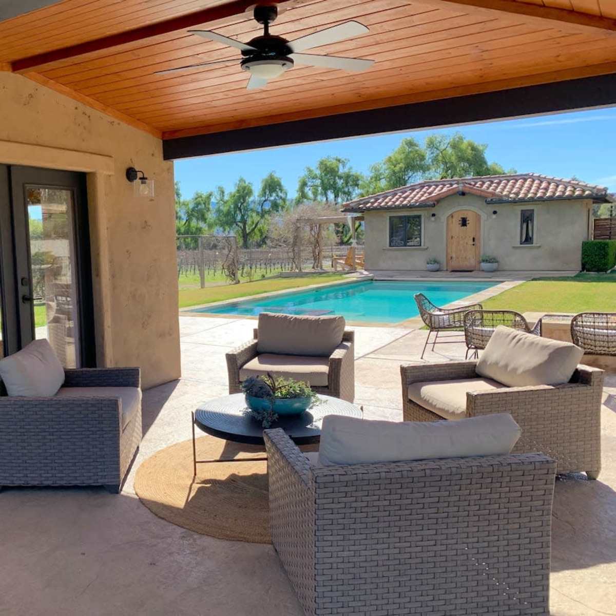 A shaded outdoor lounge area features four chairs around a circular table, adjacent to a swimming pool and a rustic building in a landscaped yard.