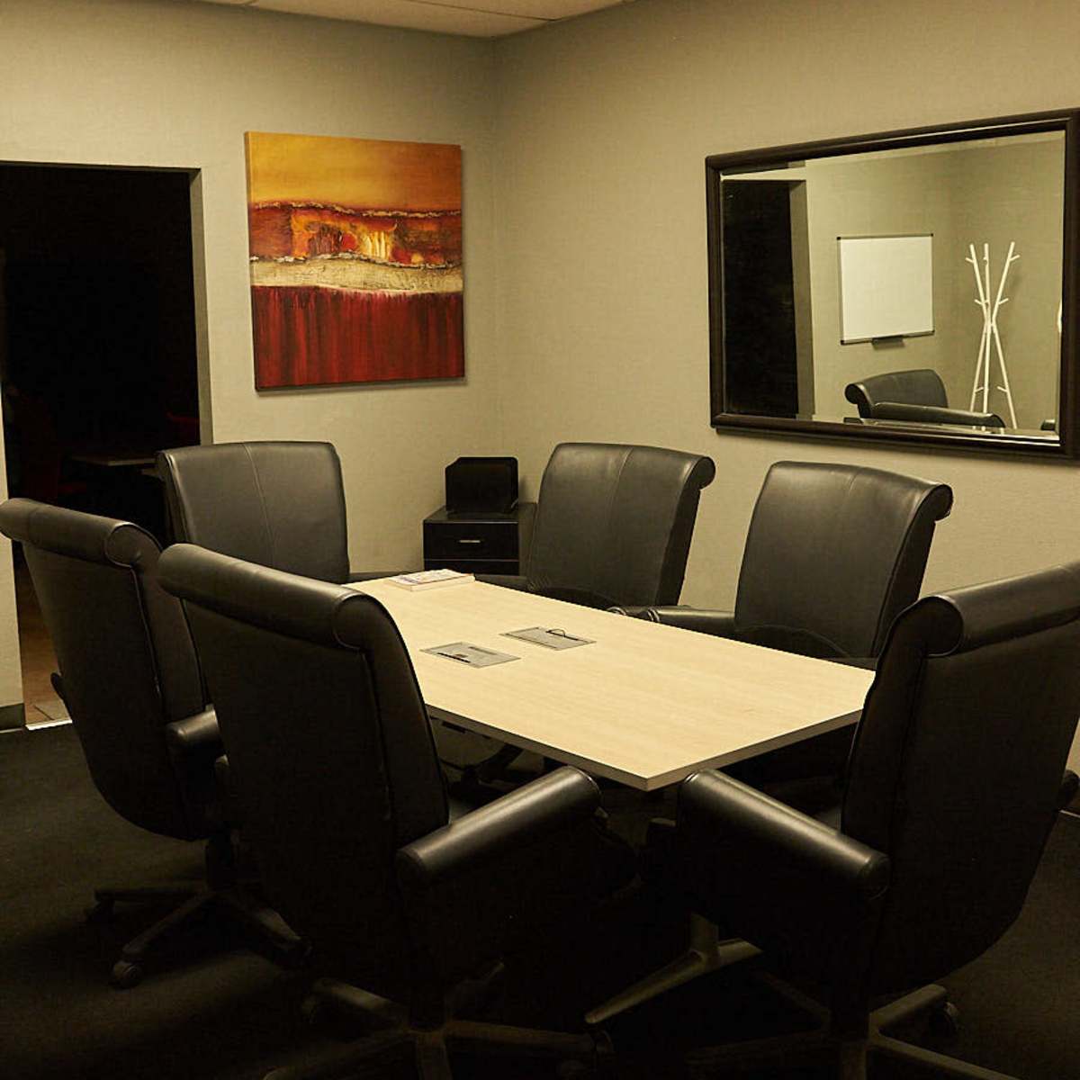 A conference room with a rectangular table surrounded by six black chairs, a large mirror, and an abstract painting on the wall.
