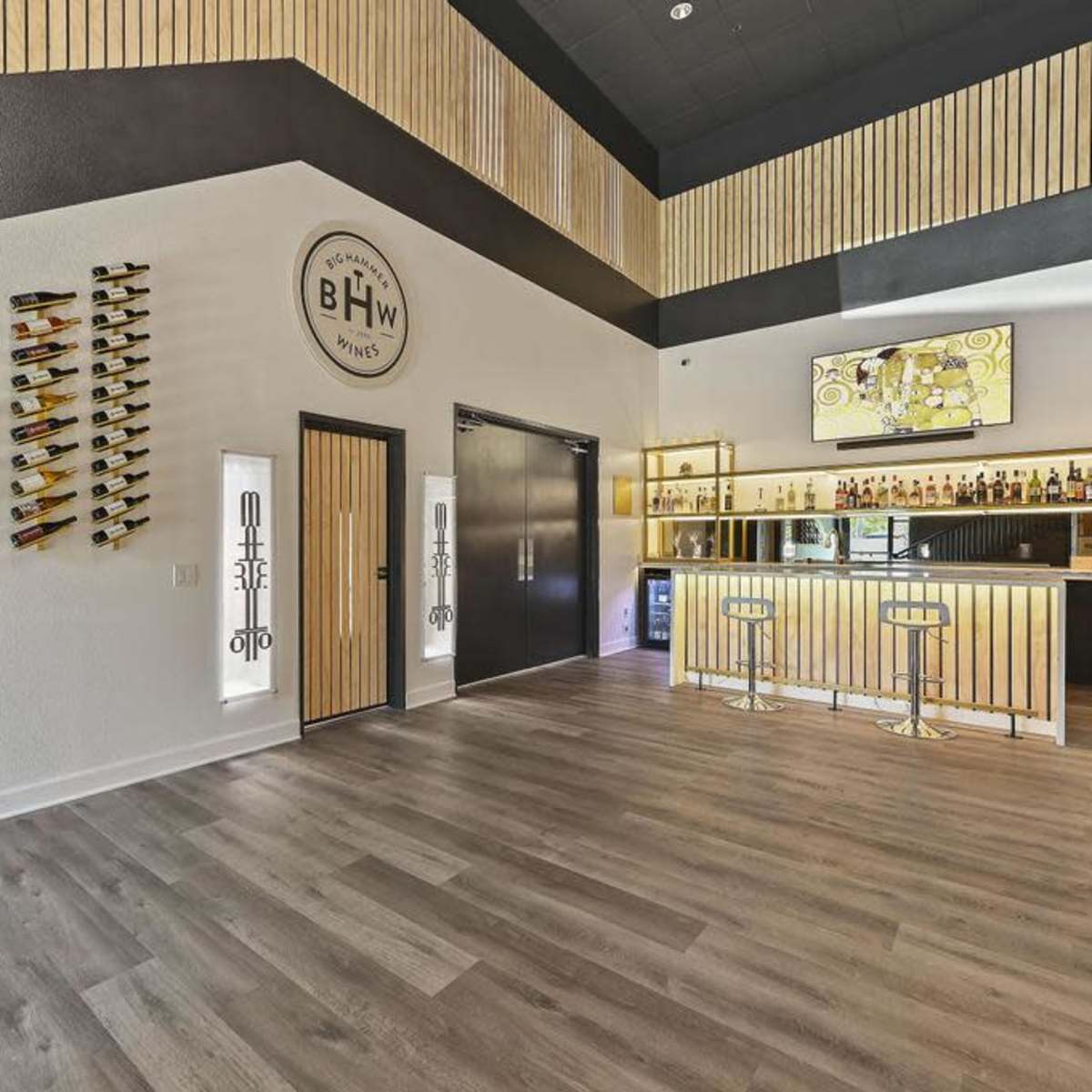 A modern bar interior featuring a wooden drink display on the wall, a well-lit bar counter, and a large television screen.