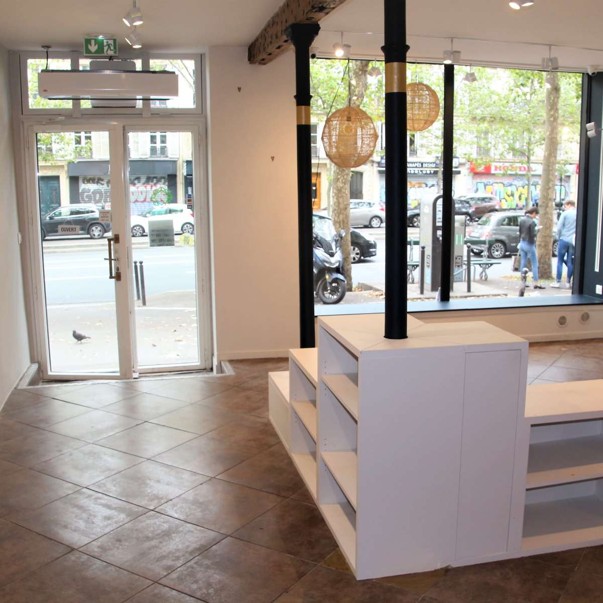 An empty room with a tiled floor, a large window displaying a street view, and a white shelving unit in the center.
