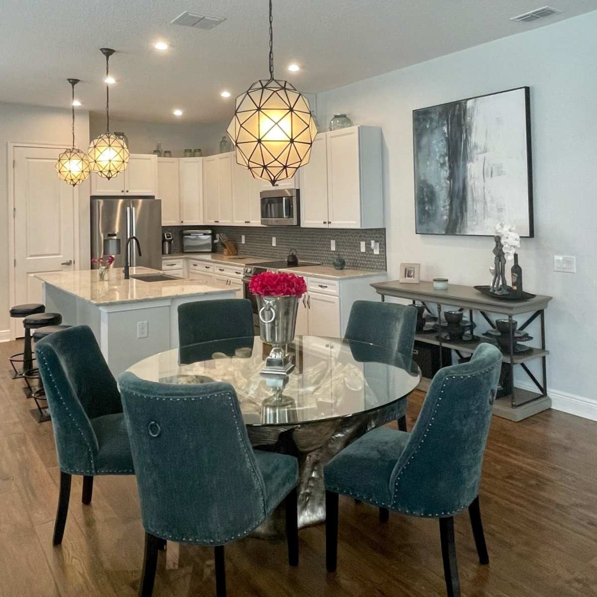 A modern kitchen and dining area with a round glass table surrounded by velvet chairs, pendant lights hanging overhead, and a stainless steel refrigerator in the background.