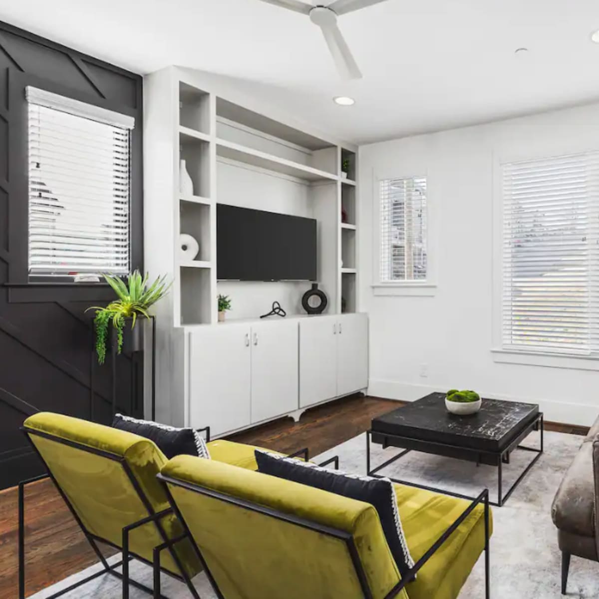 A modern living room featuring a dark accent wall, a television on a white shelving unit, a gray sofa, and two yellow chairs around a coffee table.