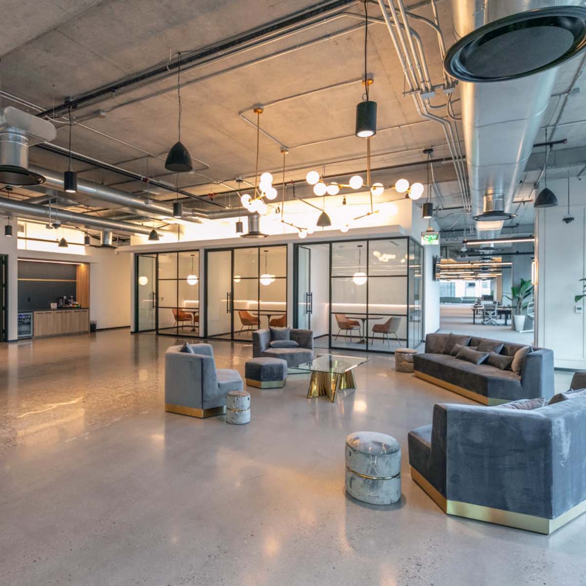 A modern office space features a seating area with gray sofas and decorative chairs, surrounded by large windows and stylish light fixtures.