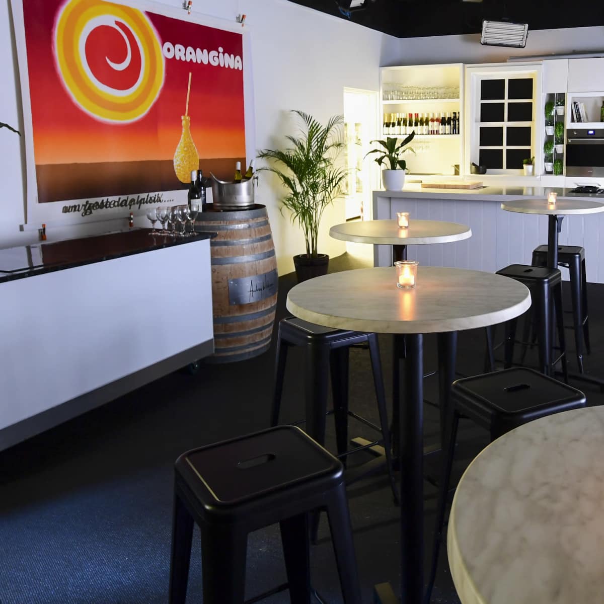 A modern bar area with round marble-top tables, black stools, and a brightly colored mural on the wall, illuminated by candles on the tables.