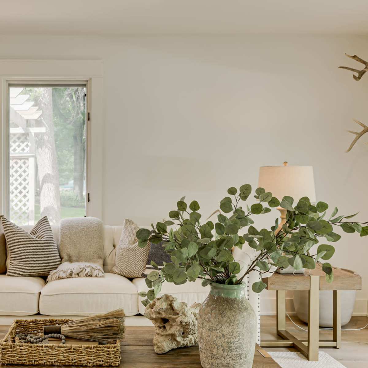 A cozy living room with a light-colored sofa adorned with various cushions, a wooden coffee table, and plants beside two lamps.