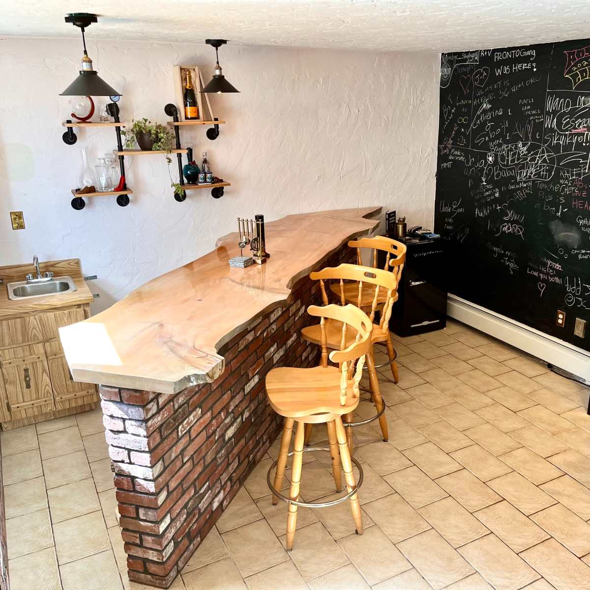 A home bar area featuring a wooden counter with three high stools, a sink to the left, a black chalkboard wall covered in writing, and shelves displaying various bottles.